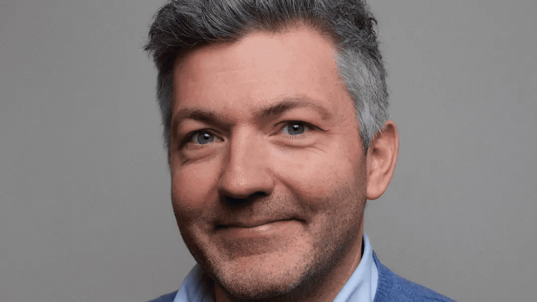 En smilende mann med kort gråaktig hår, iført blå skjorte og grå genser, poserer mot en grå bakgrunn som subtilt antyder kraften til hurtigladere for tunge kjøretøy.