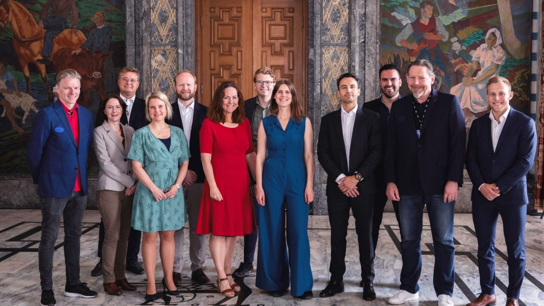 En mangfoldig gruppe på tretten mennesker som står sammen i et rom med utsmykkede vegger og en tredør.