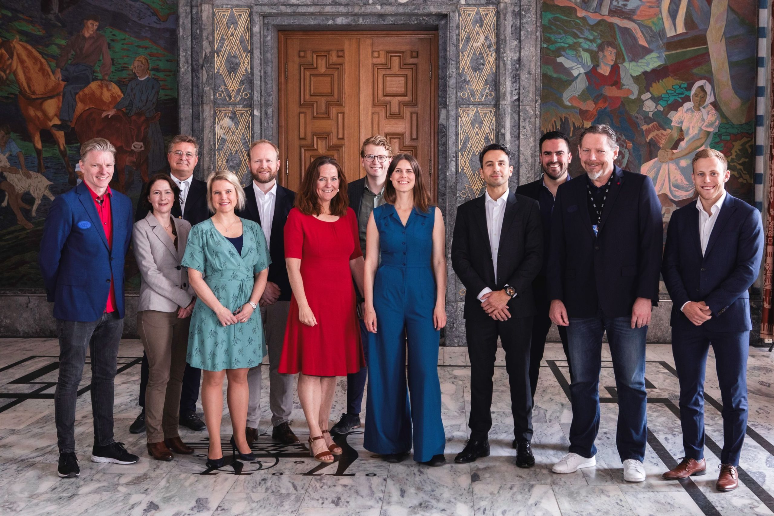 En gruppe på tolv personer står innendørs på et marmorgulv, med fargerike veggmalerier og en tredør i bakgrunnen.