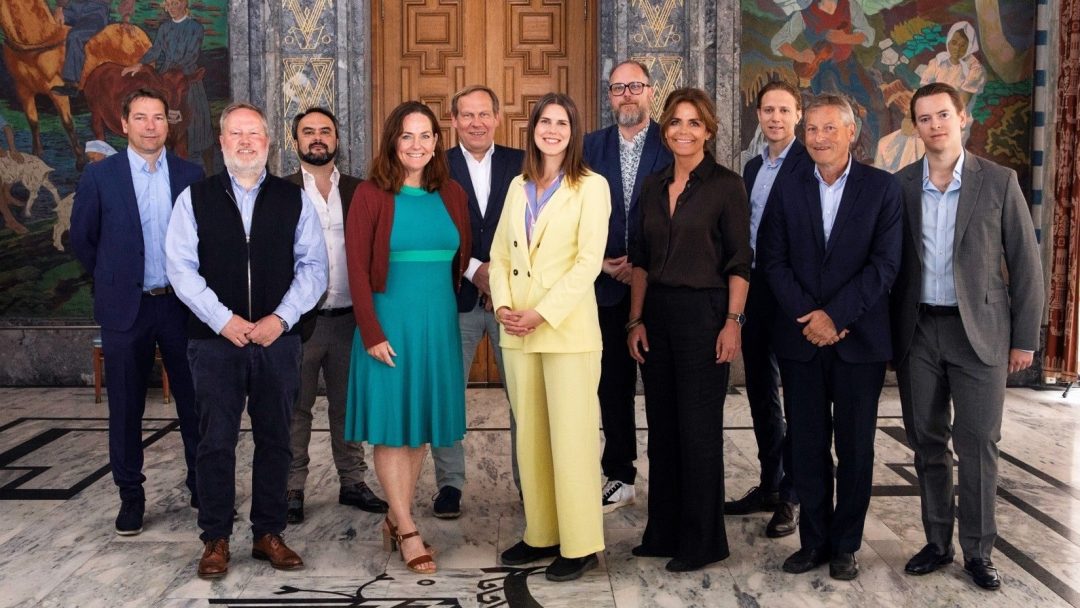 En gruppe på tolv personer, inkludert menn og kvinner, står sammen innendørs på et mønstret marmorgulv med et veggmaleri i bakgrunnen.