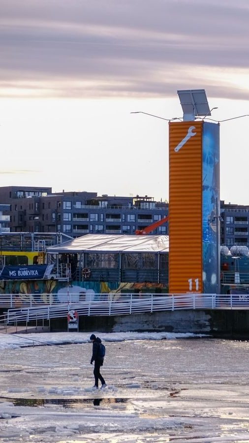 En person som rusler på den iskalde vannkanten er innrammet av en lys oransje struktur og elegante moderne bygninger, og fanger et typisk Oslotips-øyeblikk.