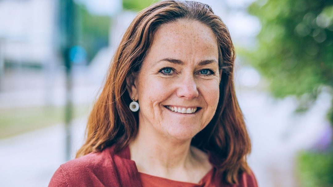 En kvinne med langt rødt hår smiler varmt, og står utendørs i et uskarpt bakteppe av frodig grønt og en svingete sti, og legemliggjør den rolige skjønnheten vi streber etter å bevare mot klimaendringers utfordringer.