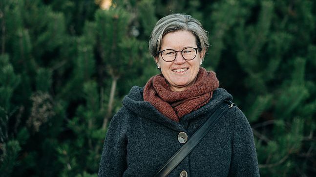 En person med kort grått hår og briller, iført mørk frakk og brunt skjerf, står og smiler midt i frodig grønt løvverk, og legemliggjør essensen av dyrking.