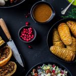 Et oppslag av juleretter uten kjøtt pryder bordet, med brødskiver, en kniv, en bolle hasselbackpoteter, spreke granateplefrø, en frisk salat og diverse sauser, alt elegant pyntet med grønt.