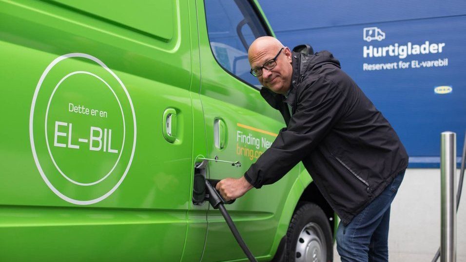 En person kobler en ladekabel til en grønn elektrisk varebil. Varebilen har norsk tekst som indikerer at det er et elektrisk kjøretøy. Et skilt i bakgrunnen nevner Hurtiglader.