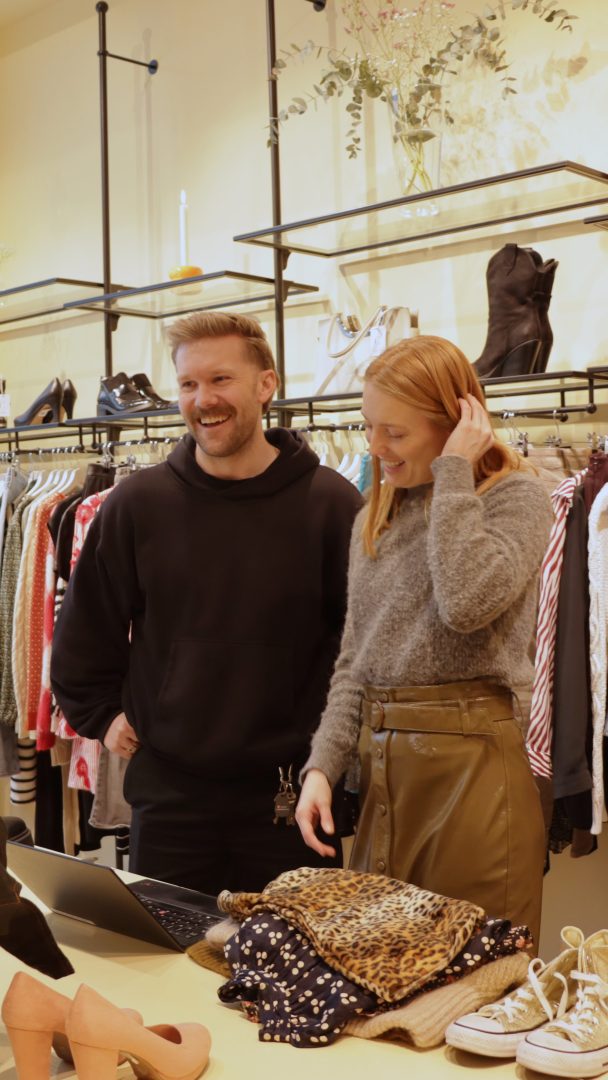 To personer som står i en bruktbutikk og smiler mens de surfer på en bærbar datamaskin. Stativ med klær og sko er synlige i bakgrunnen, og tilbyr en rekke vintagefunn.