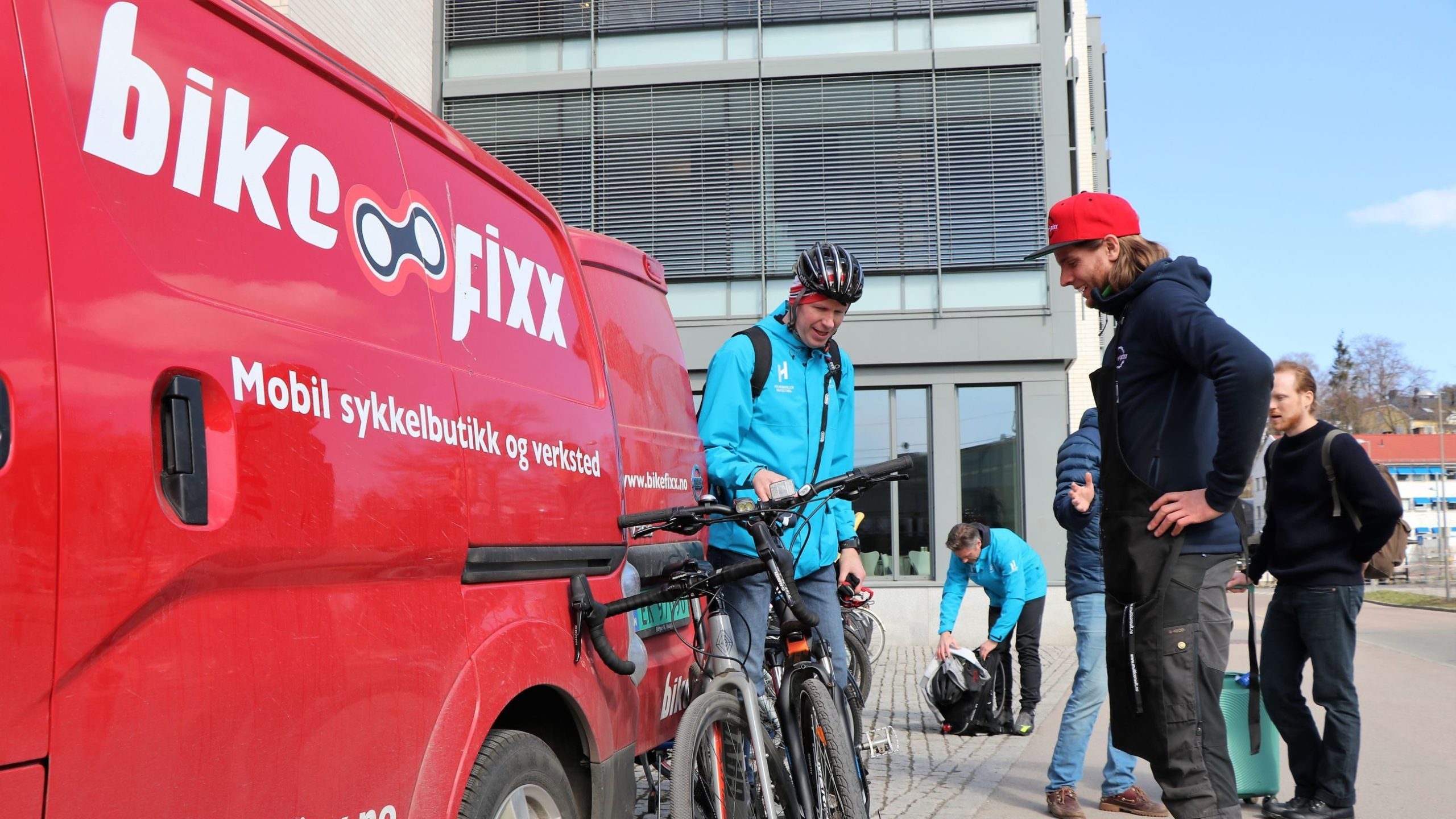 Folk som står ved siden av en rød bike fixx varebil med sykler. En person bruker hjelm, andre laster av og fikser sykler. En bygning er i bakgrunnen.