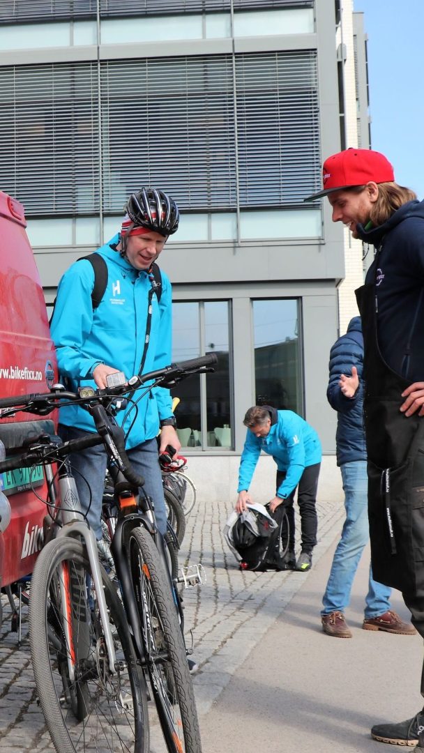 To personer i blå jakker undersøker sykler i nærheten av en rød varebil. En annen person huker seg med utstyr i bakgrunnen. En bygning med store vinduer er i bakgrunnen.