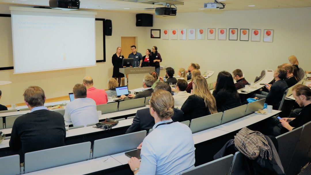 Folk som sitter i en forelesningssal og lytter til en presentasjon. Forelesere står foran ved en skjerm som viser lysbilder. De fleste deltakerne bruker bærbare datamaskiner eller tar notater.