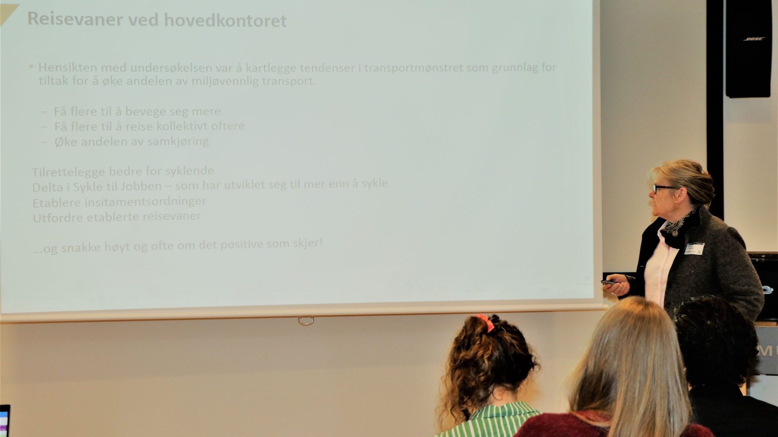 En kvinne står ved siden av en skjerm og viser en presentasjon på norsk om forbedring av transportmetoder for å øke miljømessig bærekraft. Publikumsmedlemmer sitter foran henne.