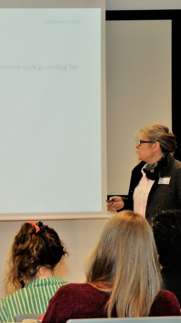 En person i en blazer står foran et projektorlerret og presenterer for et publikum.