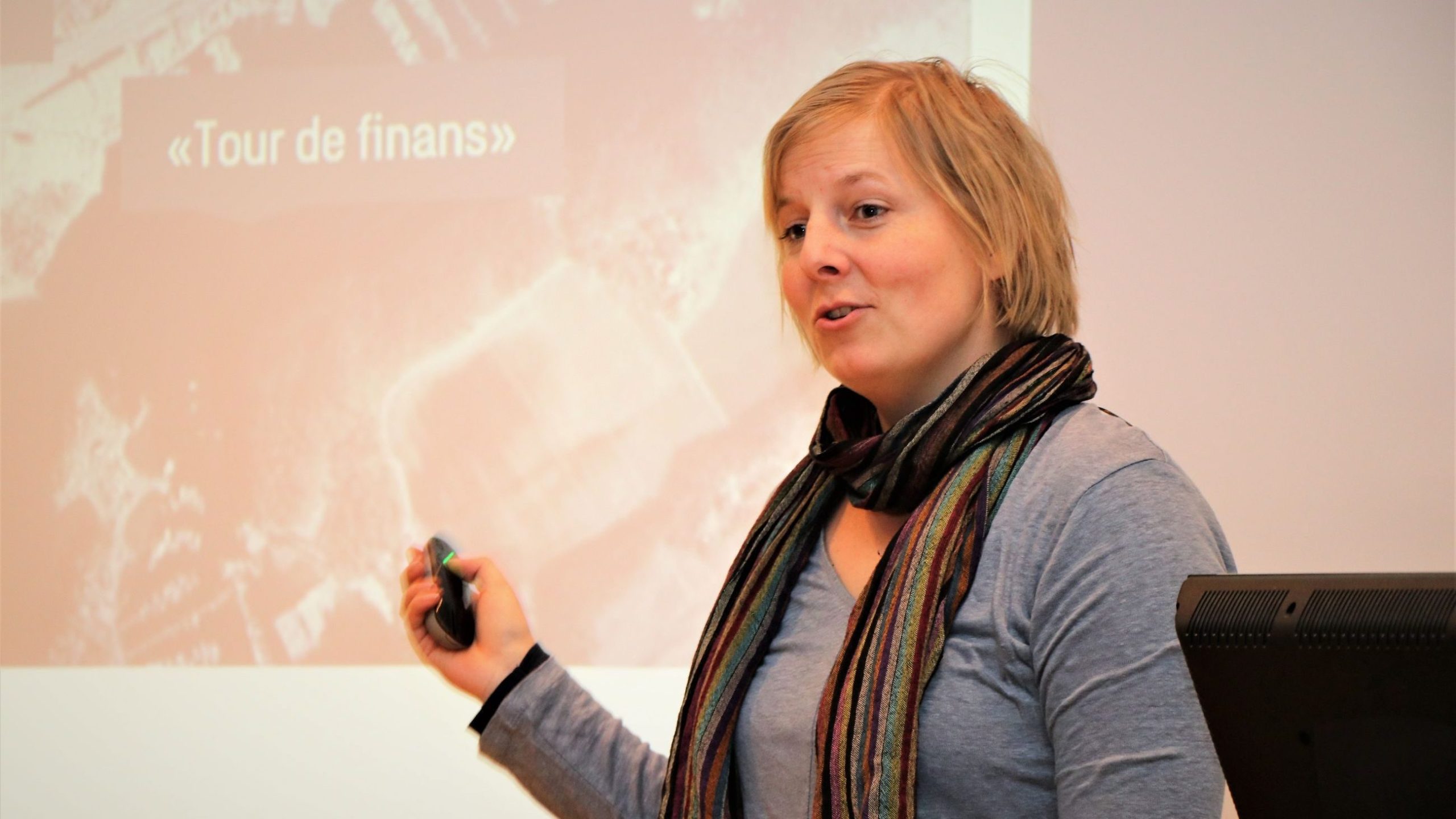 En person som gir en presentasjon med tittelen Tour de finans, holder en klikker, iført et stripete skjerf og en blå topp.