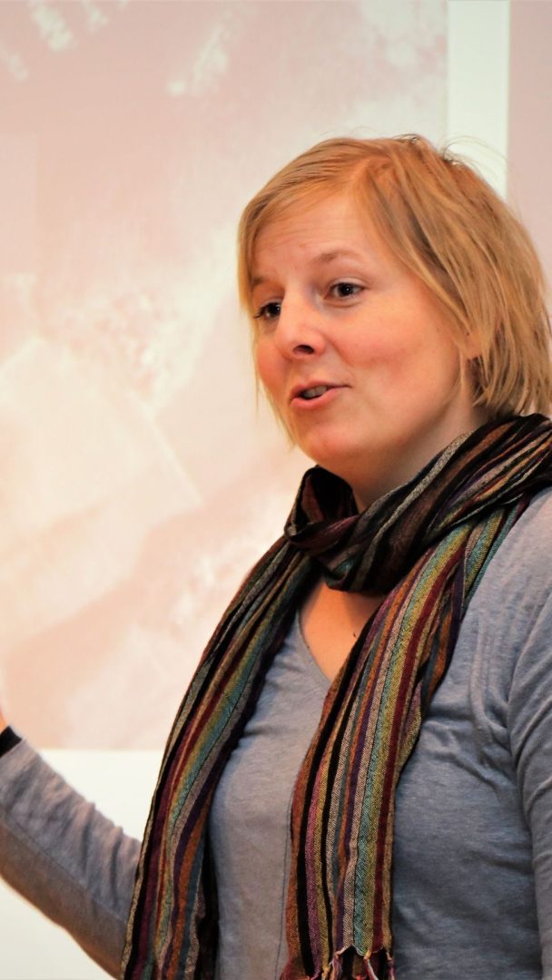 Person iført et stripet skjerf og blå topp, snakker foran en uskarp projektorskjerm.