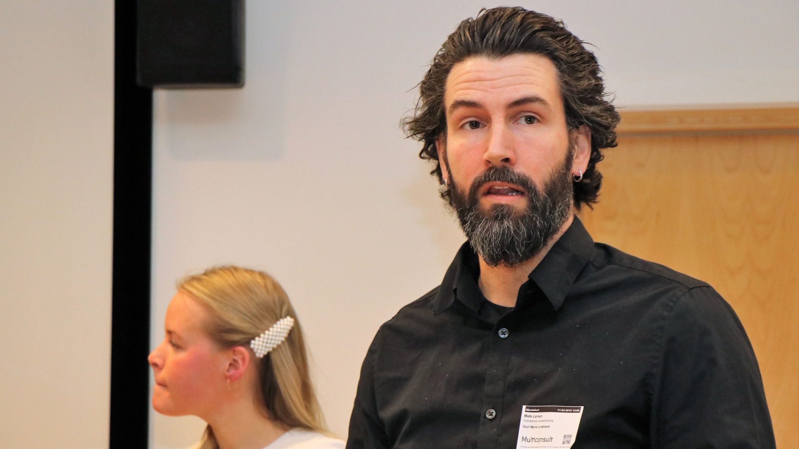 En mann med skjegg iført svart skjorte står i forgrunnen. En kvinne med en hårspenne sitter i bakgrunnen.