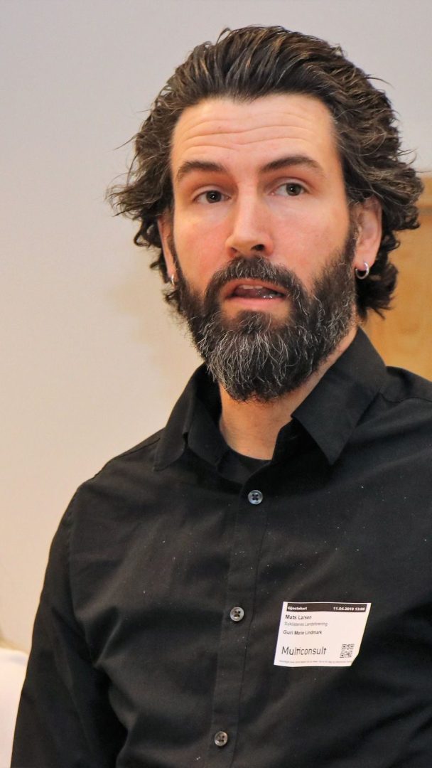 En mann med skjegg og øredobber, ikledd en svart skjorte med knapper med navneskilt, står innendørs.