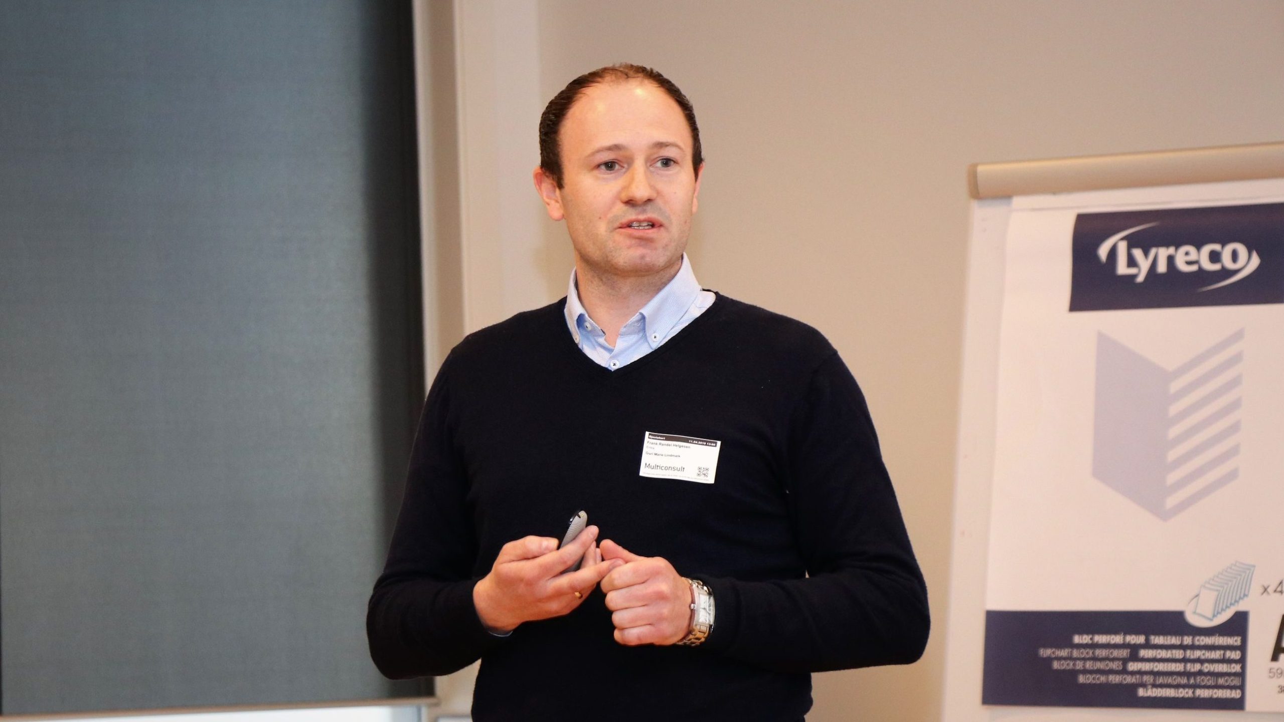 En person iført en blå genser står foran en presentasjonstavle med en Lyreco-logo og holder en peker.