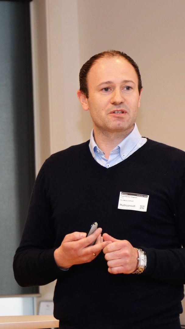 Mann i mørk genser med navneskilt, holder en presentasjonsfjernkontroll, snakker i et rom.