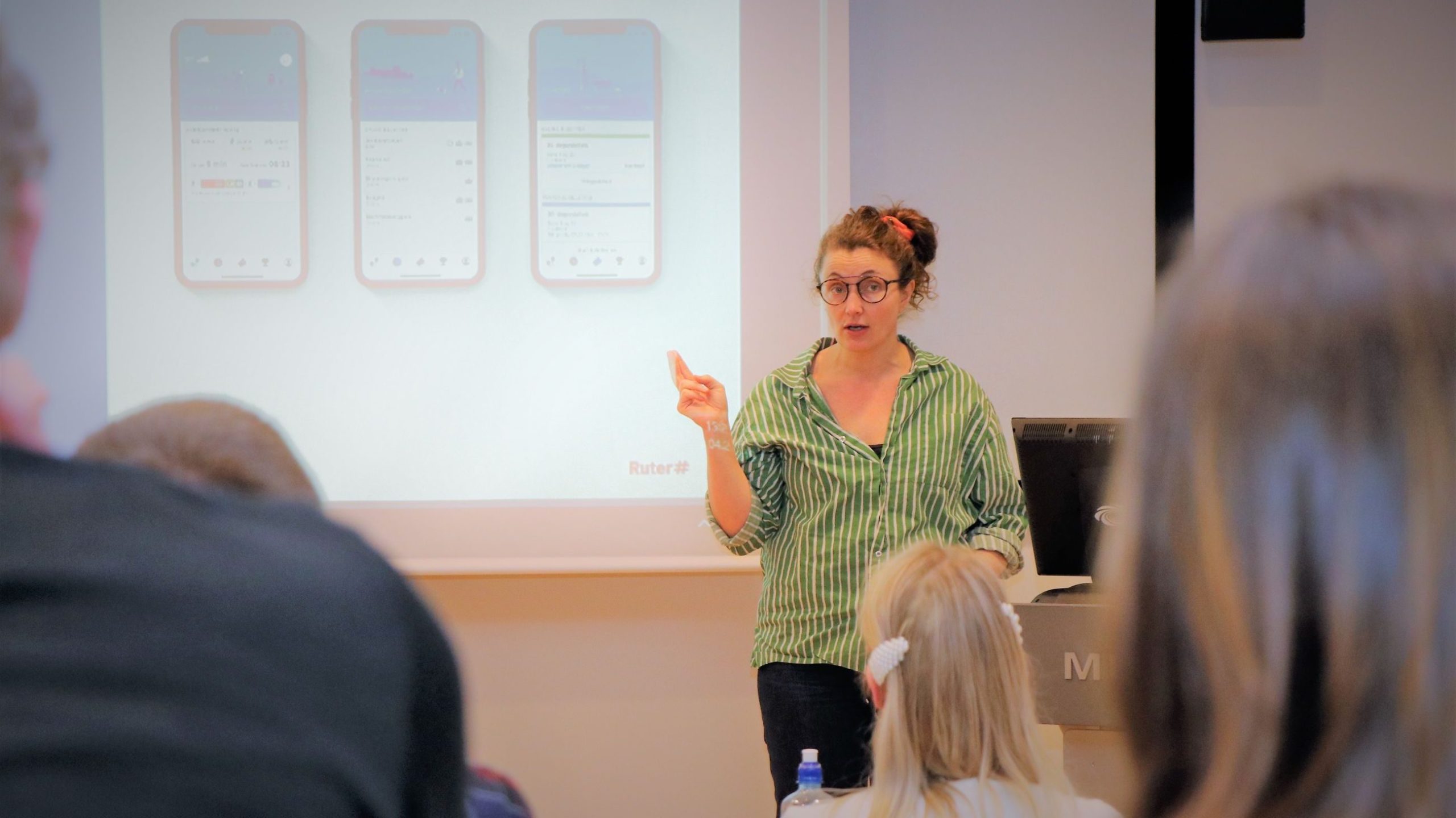 En kvinne i stripet skjorte holder en presentasjon om smarttelefonoppsett, og peker på en skjerm som viser app-grensesnitt, foran et publikum.