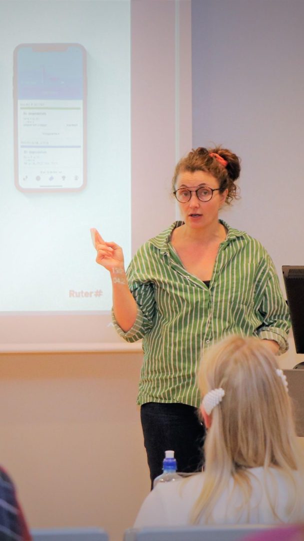 En kvinne i en grønnstripet skjorte presenterer seg foran en skjerm som viser et mobilappgrensesnitt.