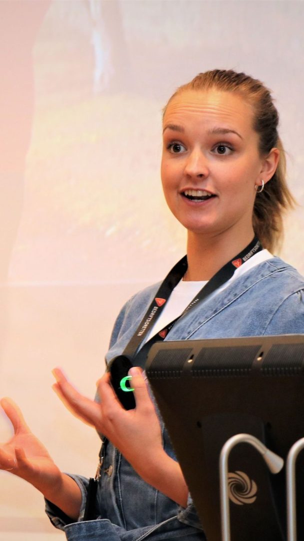 Person som presenterer og gestikulerer med begge hender, iført en snor, foran en skjerm.