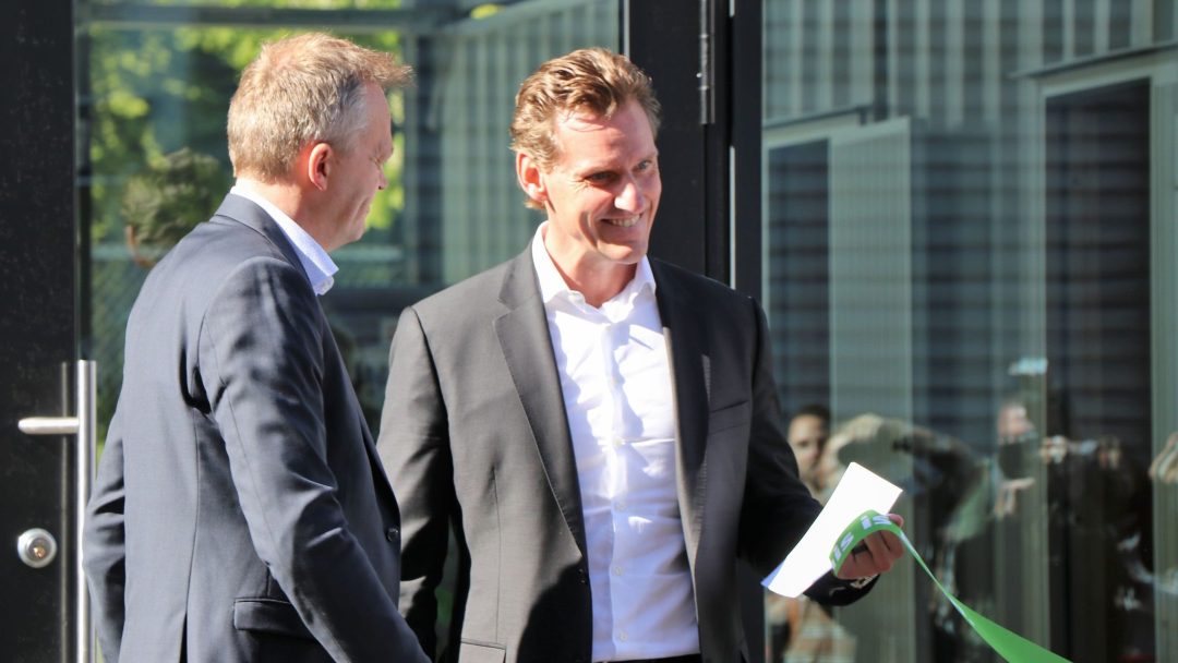 To menn i dress står nær en glassdør. Den ene holder et bånd og konvolutt, eventuelt deltar i et utendørs arrangement.