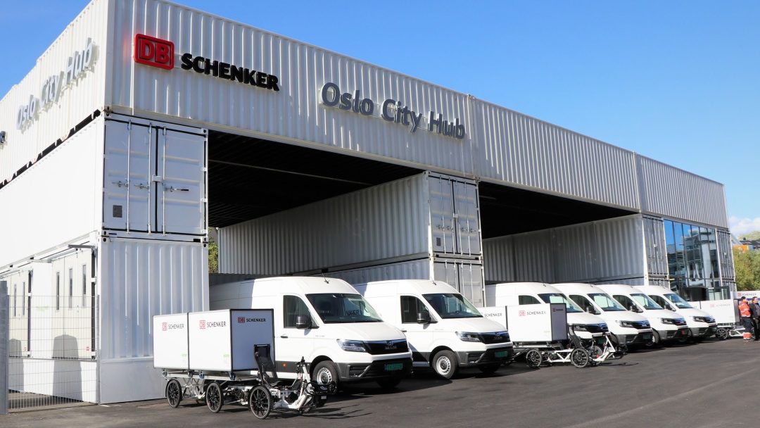 DB Schenker Oslo City Hub med flere hvite varebiler og lastesykler parkert foran. Stor containerstruktur under en klar blå himmel.
