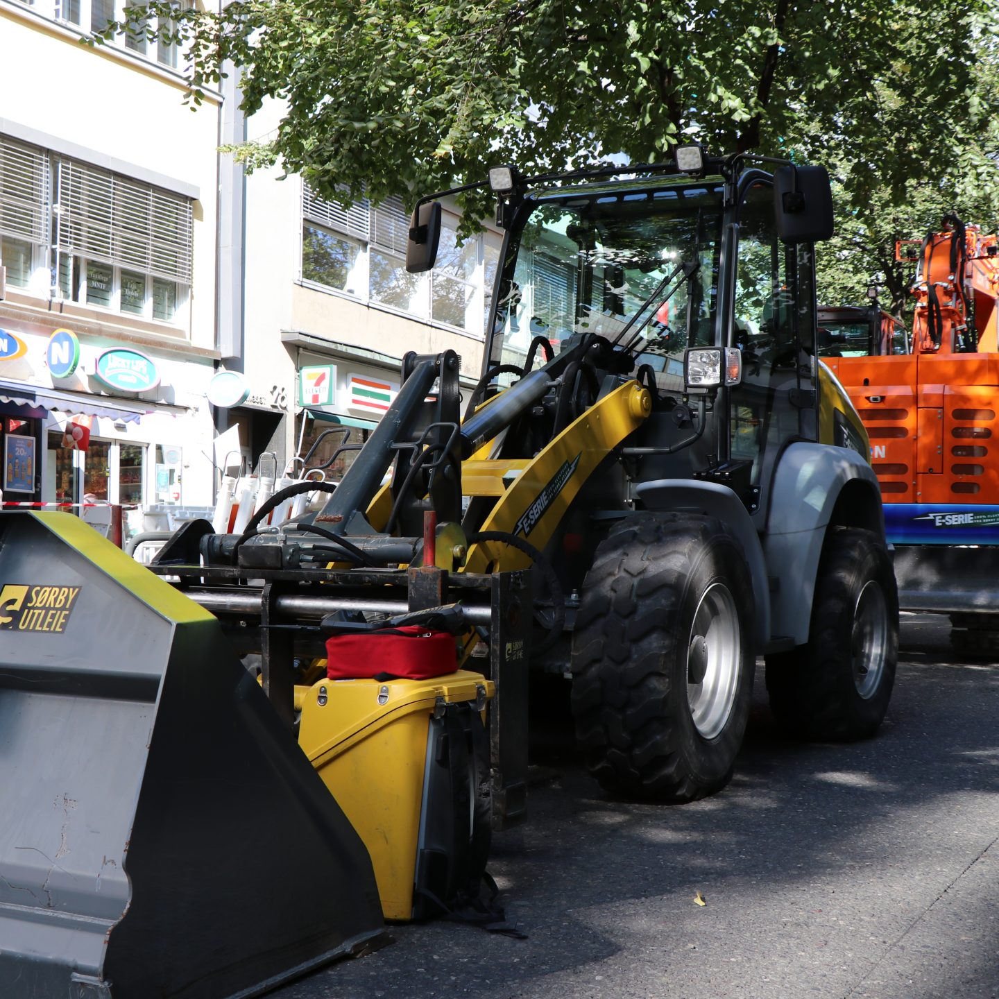 Byggekjøretøyer, inkludert en hjullaster og en gravemaskin, parkert i en bygate oppfyller klimakravene, med trær og butikker i bakgrunnen.