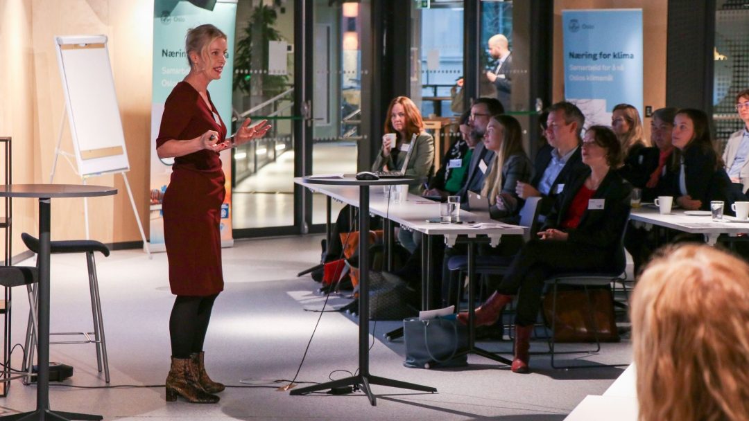 En kvinne i en rød kjole presenterer for et sittende publikum i et konferanserom. En flippover og bannere er i bakgrunnen.