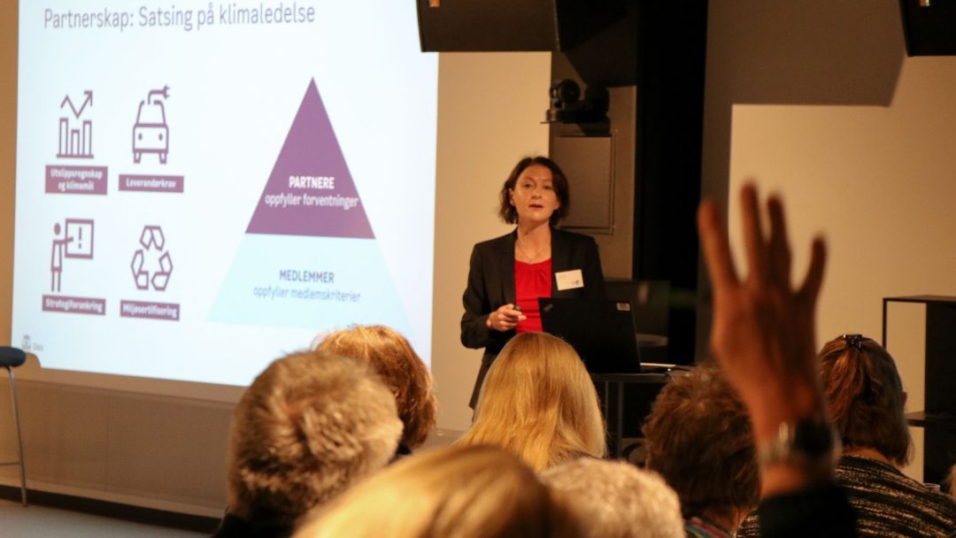 En kvinne presenterer et lysbilde om klimarelaterte partnerskap for et publikum. En person i forgrunnen rekker opp en hånd, muligens forbereder seg på å stille et spørsmål.