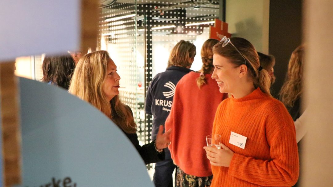 To kvinner er i samtale på et arrangement. Den ene har på seg et svart antrekk, og den andre en oransje genser og holder et glass. Andre er i bakgrunnen.