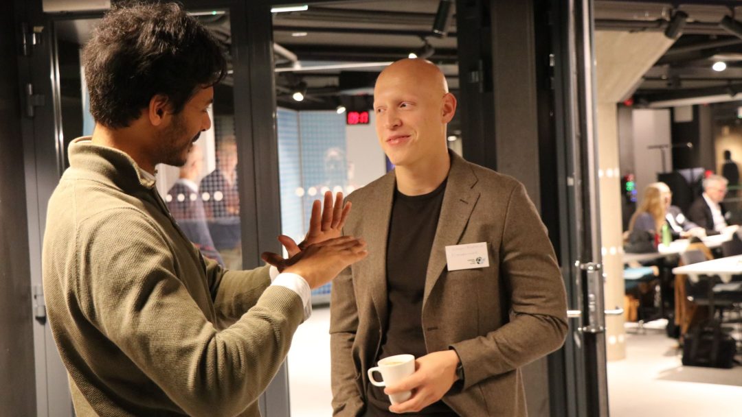 To menn står i samtale innendørs; den ene gestikulerer mens han snakker, og den andre holder en kaffekopp. Begge har på seg forretningsantrekk i moderne kontormiljø.