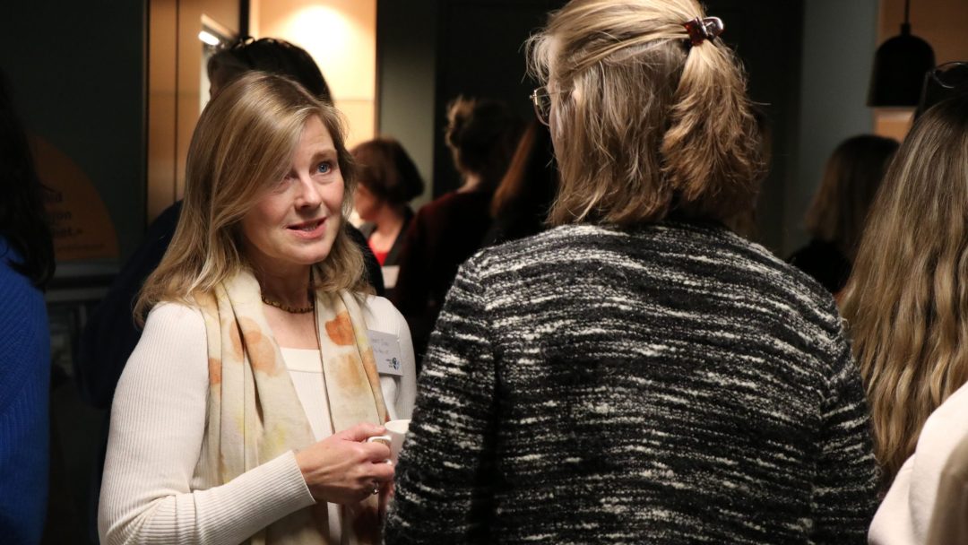 To kvinner er i samtale på en innendørs sammenkomst, med flere andre personer i bakgrunnen.