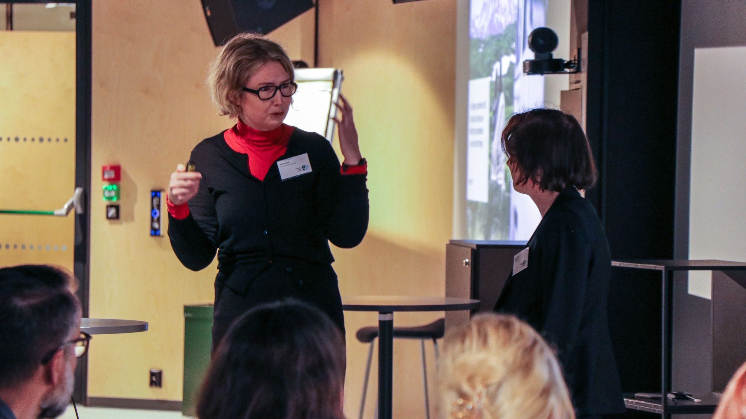 To kvinner er engasjert i samtale i en innendørs setting. Den ene snakker med en uttrykksfull gest mens den andre lytter. Folk sitter i forgrunnen.