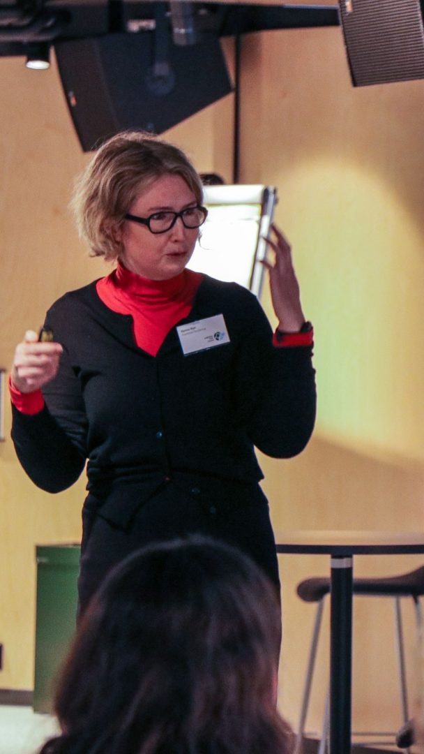 Person i briller og svart antrekk holder en presentasjon innendørs, gestikulerer med én hånd, med en publikummer synlig i forgrunnen.