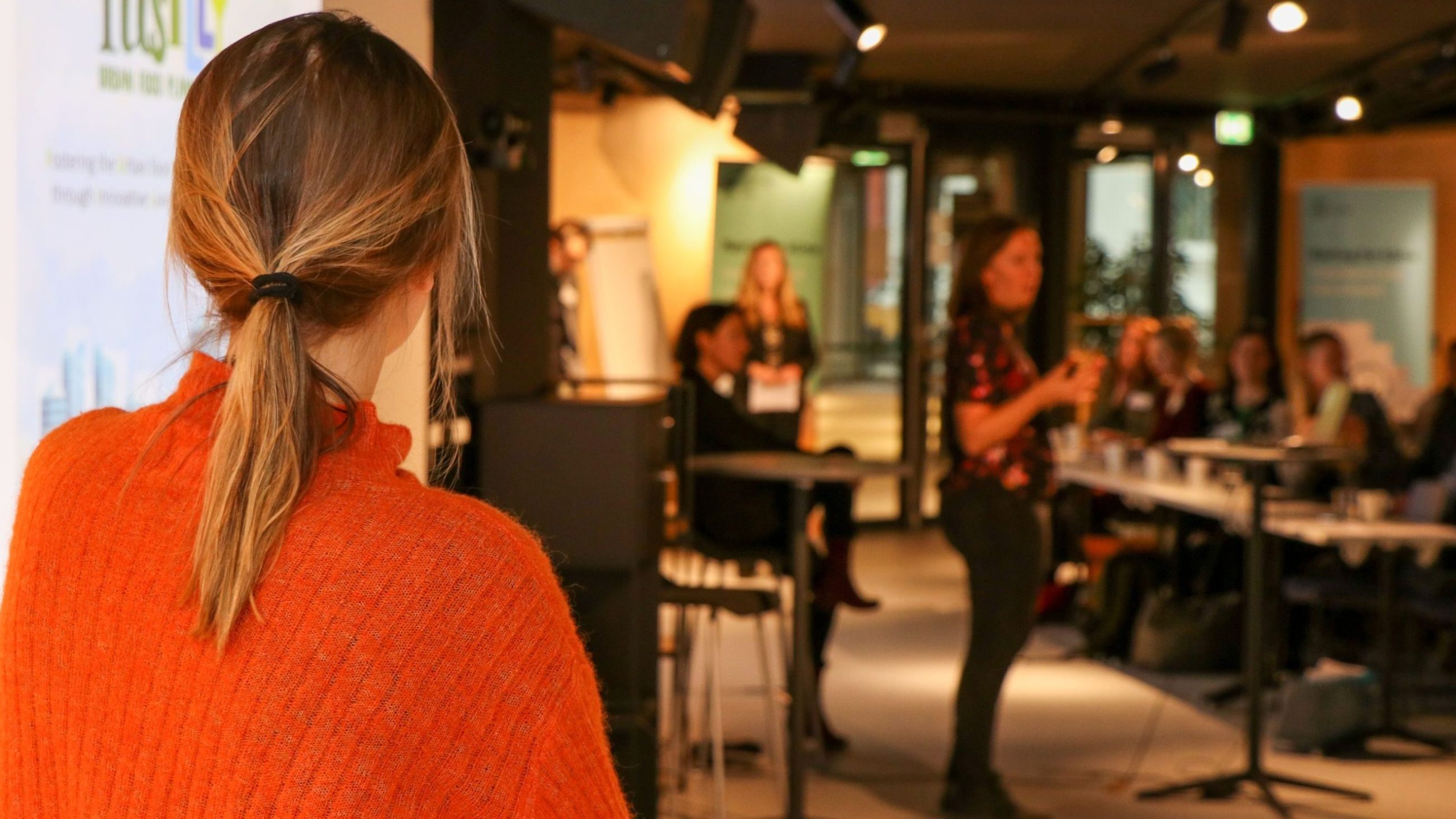 En kvinne i en oransje genser ser på en annen kvinne som snakker til et publikum i et konferanserom.