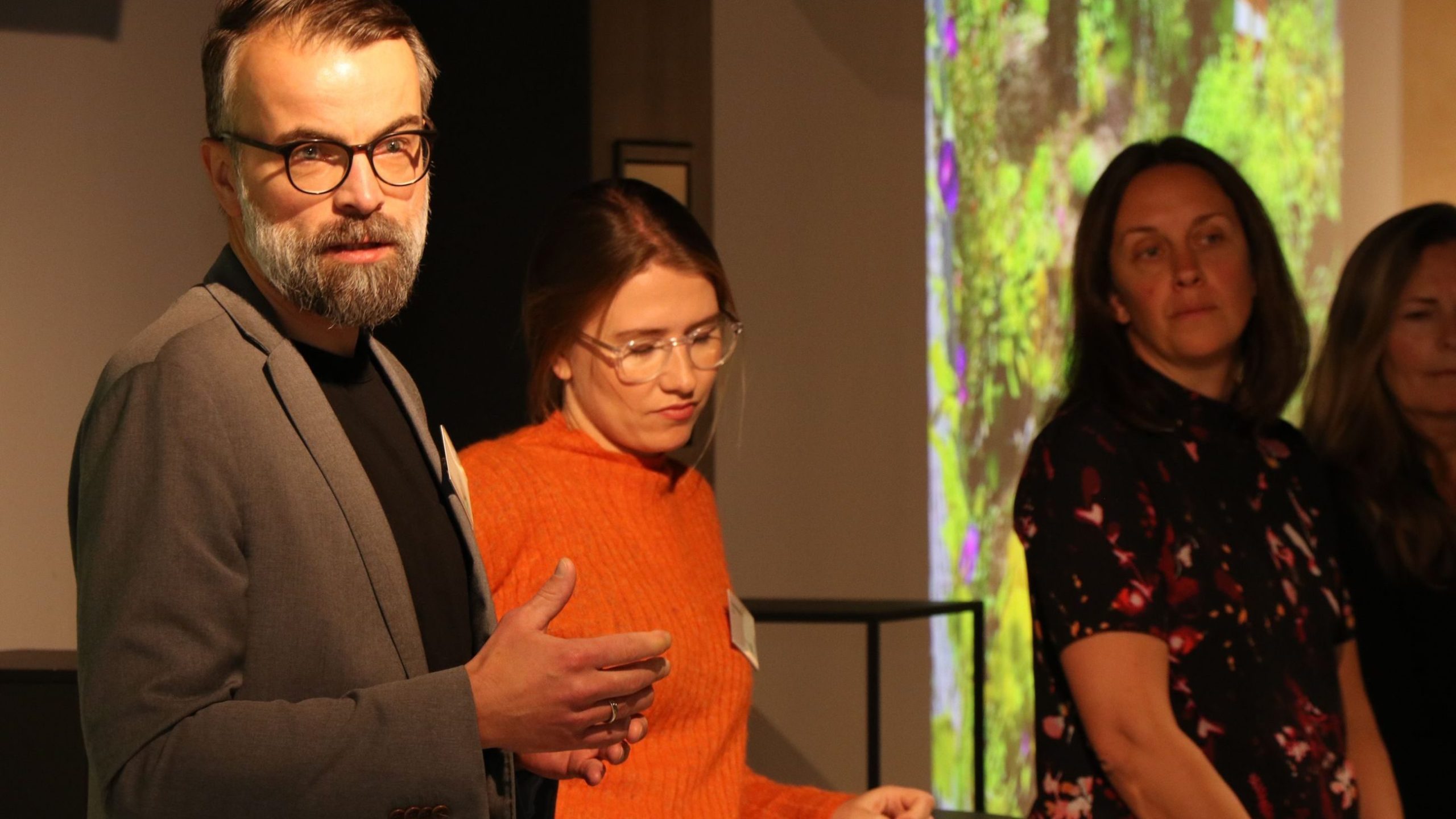 En mann i en grå blazer snakker, stående ved siden av tre kvinner. En kvinne bruker briller og en oransje genser. Et fargerikt naturbilde projiseres i bakgrunnen.