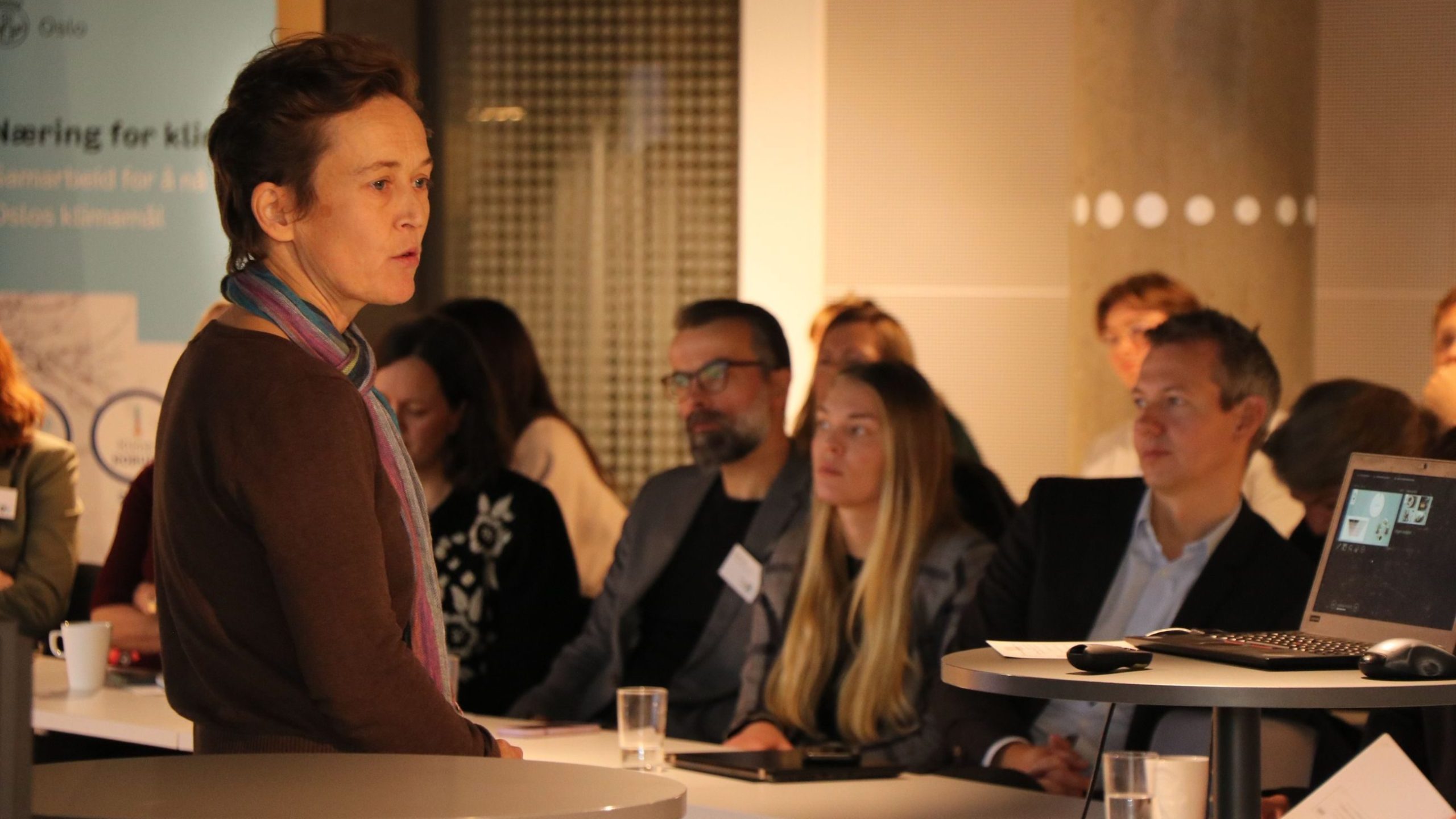 En person snakker til et publikum i et konferanserom. En bærbar datamaskin på et bord viser en presentasjon. Deltakerne sitter og er oppmerksomme.
