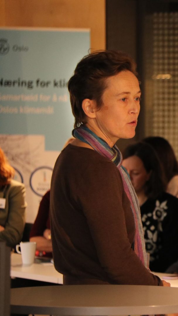 En person med kort hår og et fargerikt skjerf snakker på et innendørs arrangement. Andre sitter i bakgrunnen nær et banner.