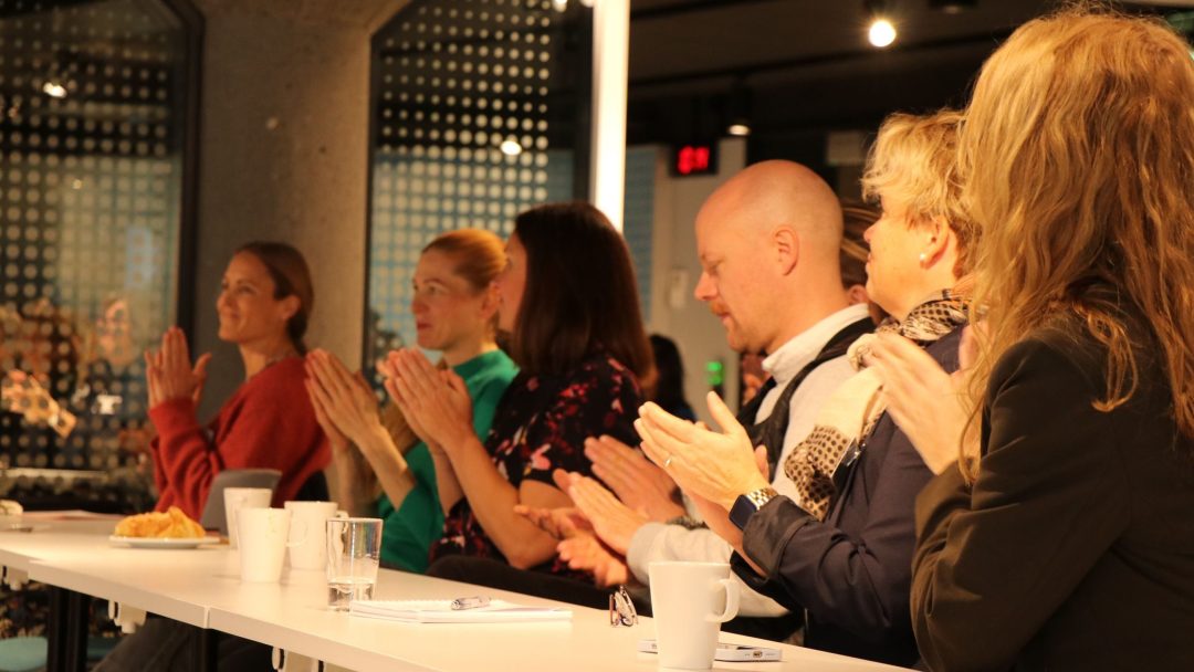 En gruppe mennesker sitter ved et hvitt bord og klapper. Noen har kopper og glass på bordet. Bakgrunnen har et moderne, prikkete design.