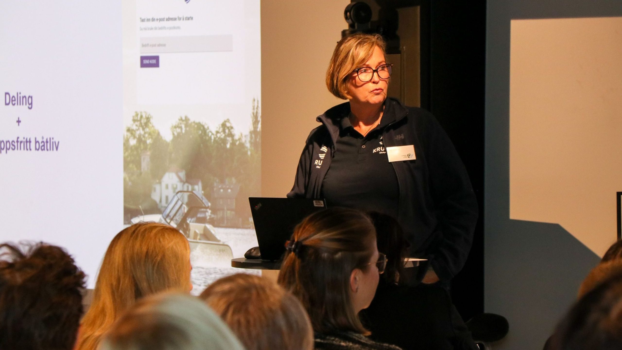 En kvinne står på et podium med en bærbar datamaskin og snakker til et sittende publikum. Et presentasjonslysbilde vises i bakgrunnen.