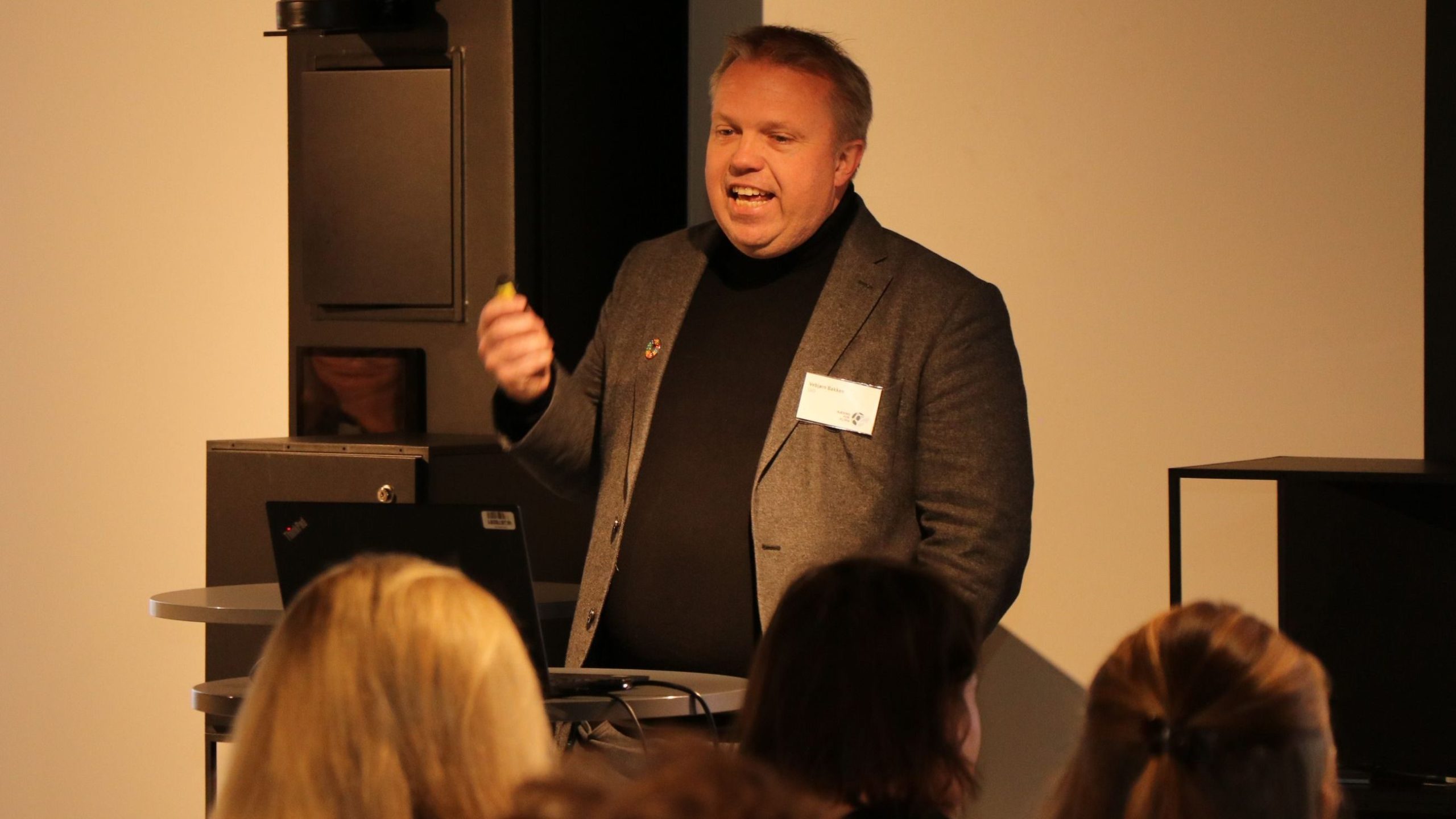 En mann i dress holder en liten gjenstand i en presentasjon. En gruppe mennesker lytter oppmerksomt foran.