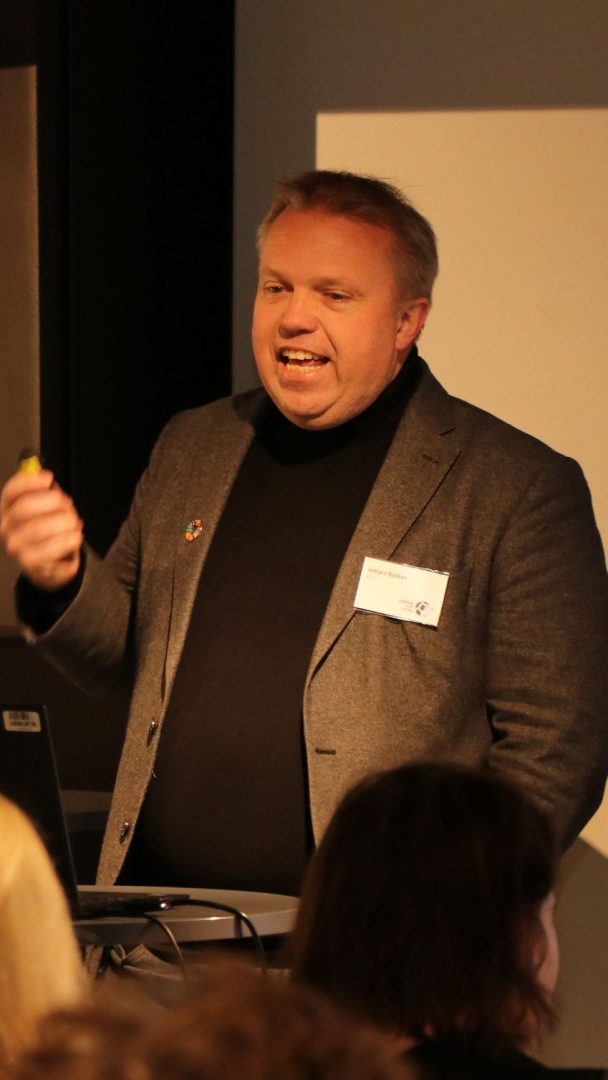 En person iført en grå blazer taler ved et podium innendørs.