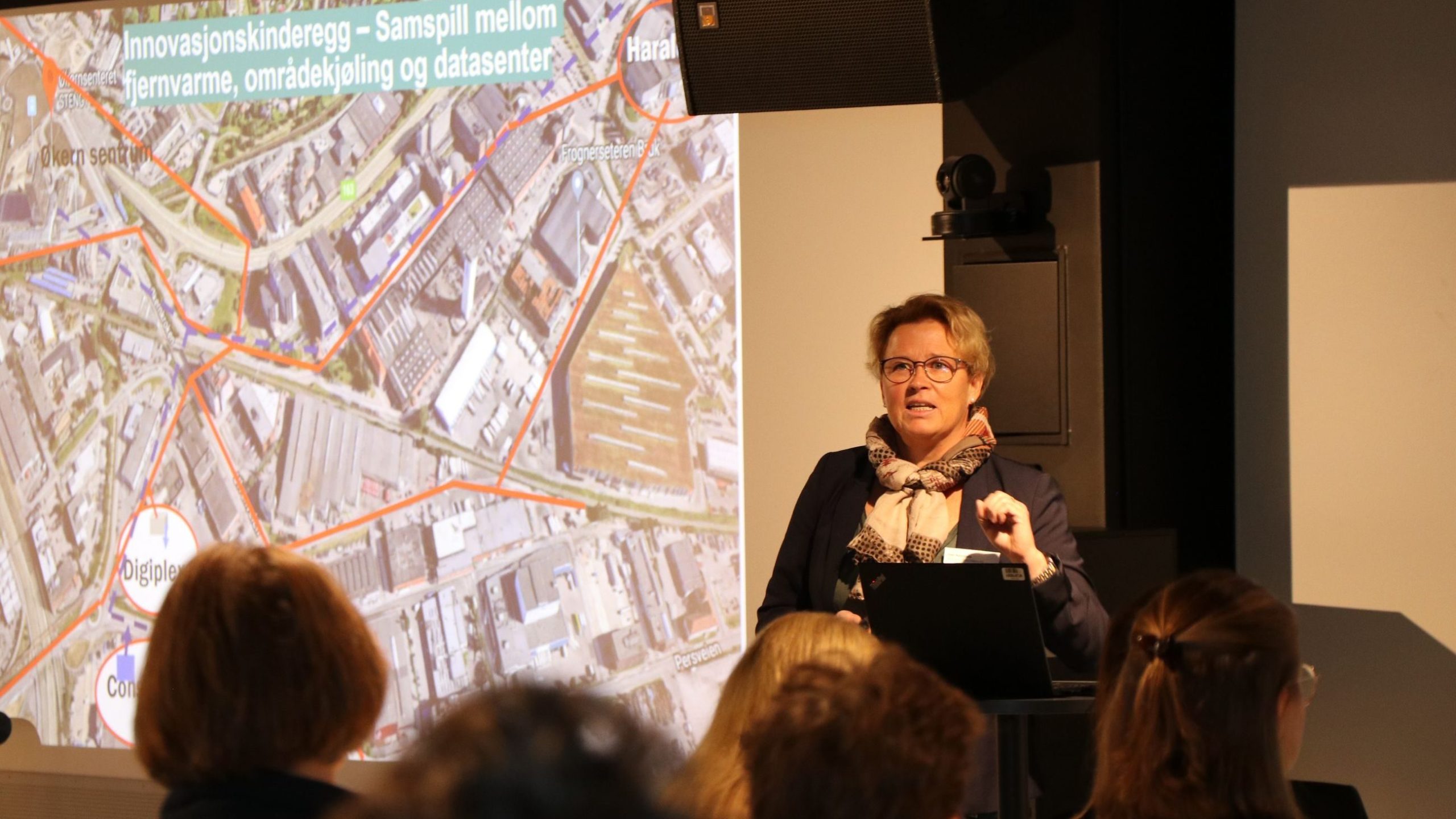 Person som presenterer på en konferanse med et kart over et byområde vist på skjermen, og fremhever fjernvarme- og datasenterplasseringer. Publikumsmedlemmer er synlige i forgrunnen.