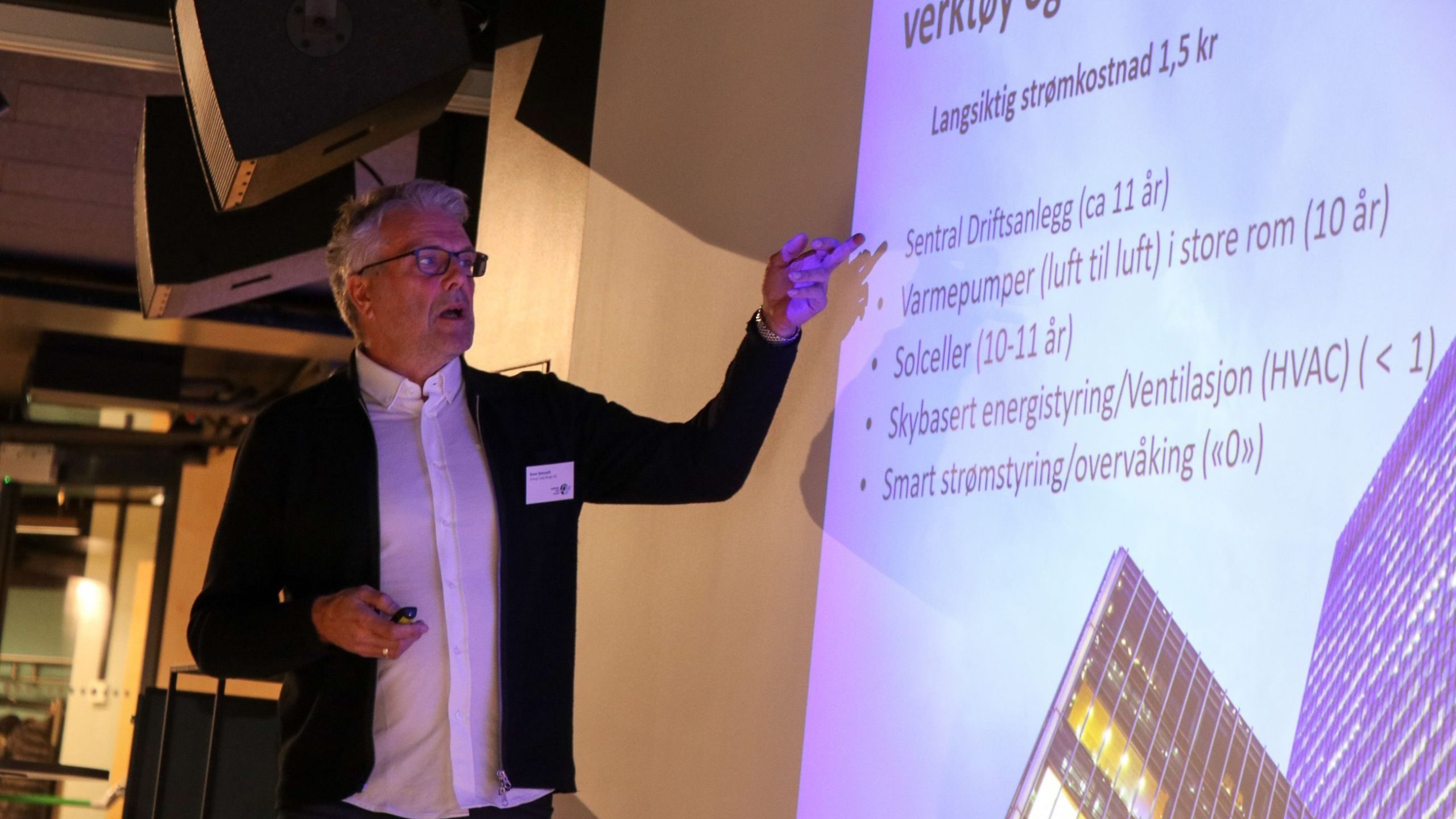 En person som holder en presentasjon og peker på et lysbilde med tekst og et byggebilde.