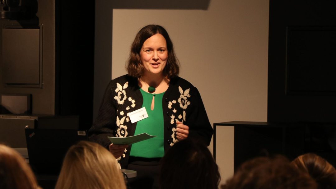 En kvinne i en svart cardigan med hvite blomstermønstre snakker til et publikum, holder et stykke papir, stående nær en bærbar PC og projektor.