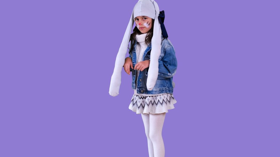 Et barn i et halloweenkostyme-kanindrakt, med lange ører og ansiktsmaling, har på seg en dongerijakke, skjørt og strømpebukser mot en lilla bakgrunn.