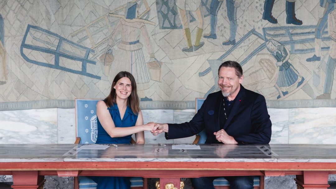 To personer satt ved et bord, smilende og nevestøtende. De står foran et stort, kunstnerisk veggmaleri.