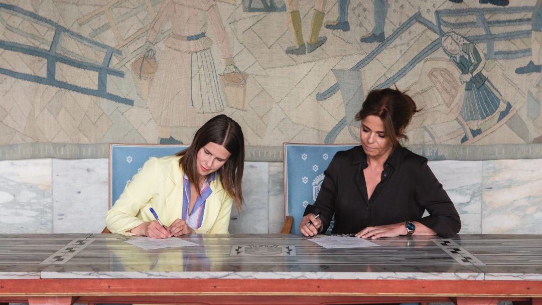 To kvinner som satt ved et bord, fokuserte på å signere dokumenter. Et detaljert veggmaleri som viser mennesker og mønstre fungerer som bakteppe.