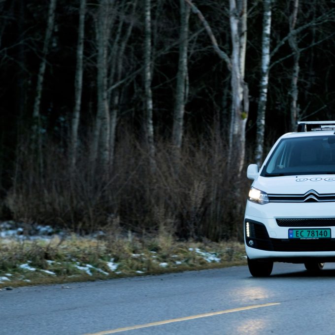 En hvit Citroën-varebil med takstativ kjører på en asfaltert vei, med nakne trær og sparsom snø i bakgrunnen.