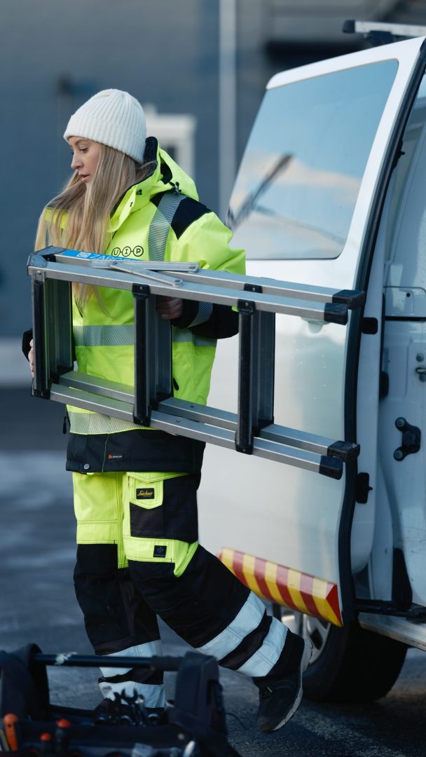Person i refleksutstyr og en lue som bærer en stige fra et kjøretøy, utendørs.